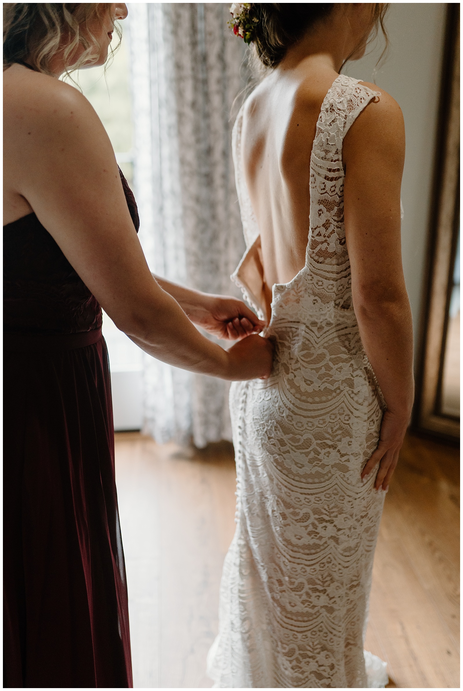 A bridesmaid helps zip up the back of a lacey fitted bridal gown.