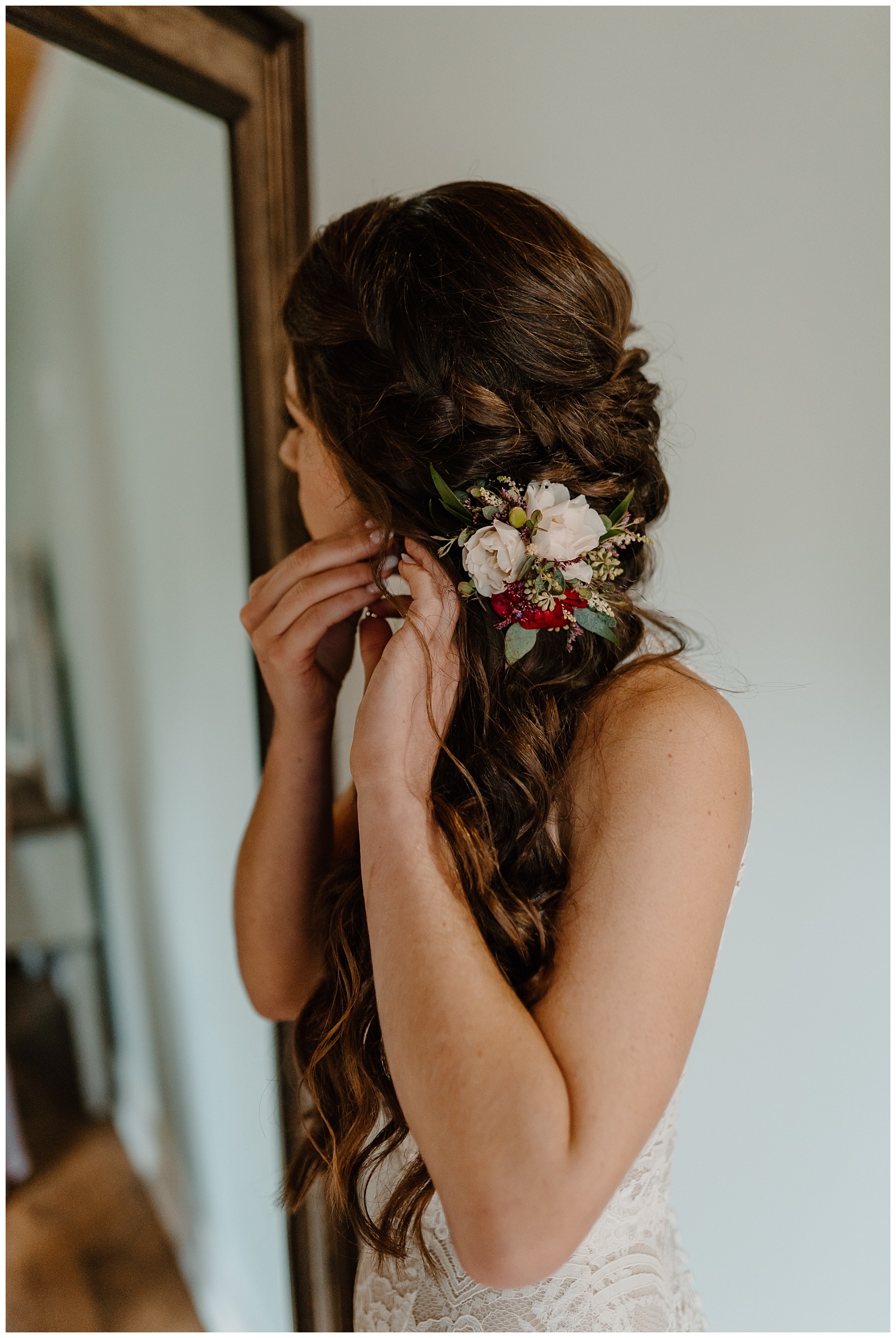 Bride with long braided hair style and matching flowers in her hair is putting on her earrings and looking in the mirror.