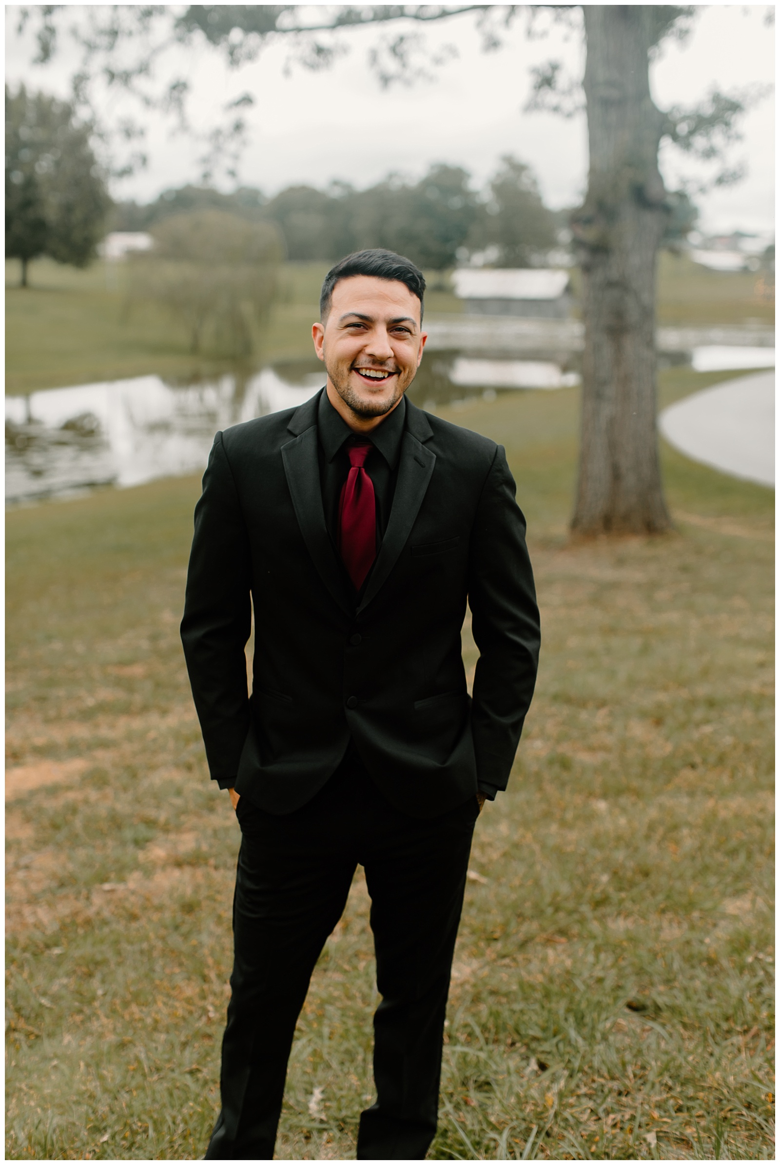 Portrait of a Groom outside by a small pond before his wedding. He is wearing a black tailored suit, black shirt and pants, and a dark, bold bordeaux tie.