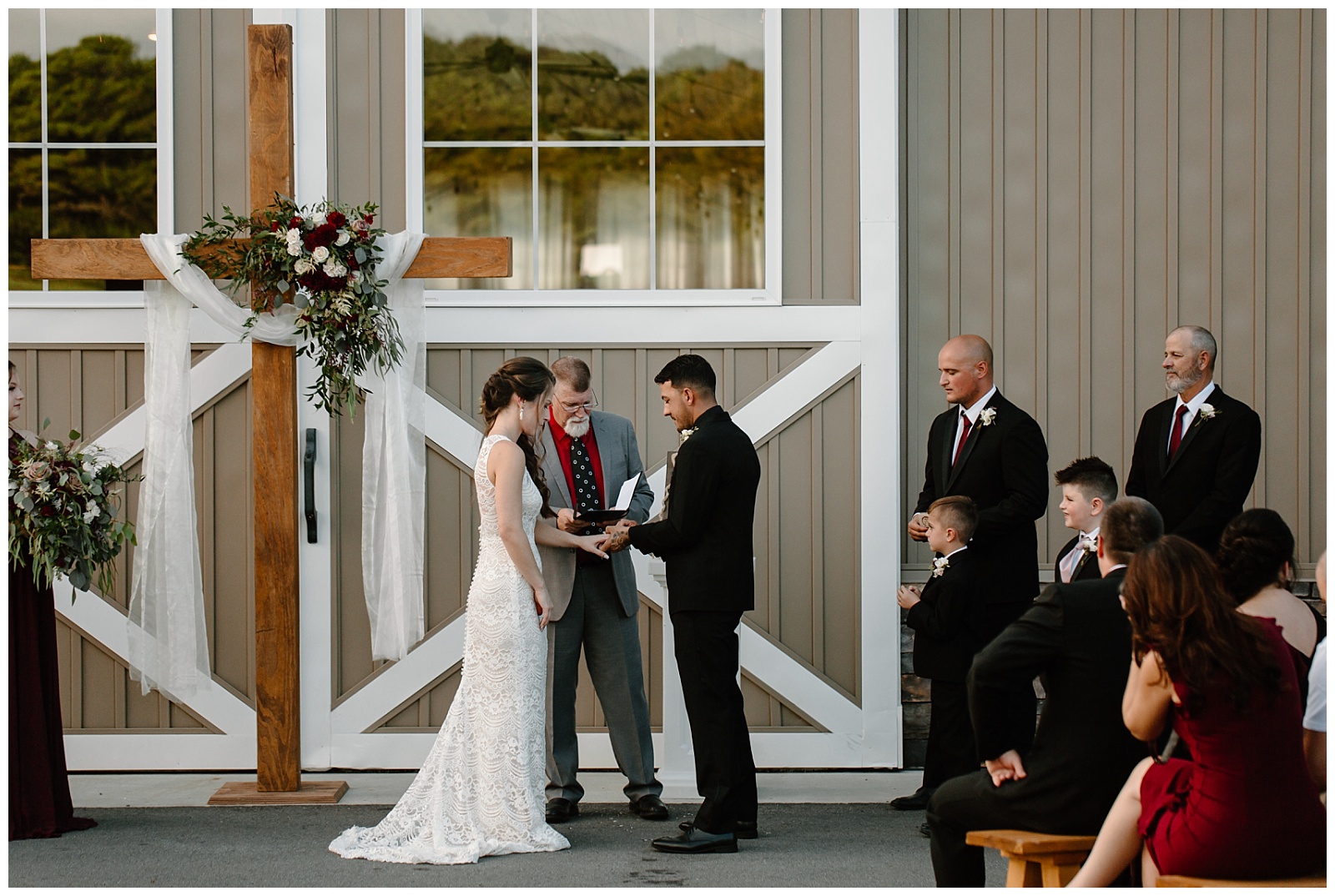 Bride and groom stand hand in hand and place rings on each other's fingers during outdoor wedding ceremony in front of barn doors