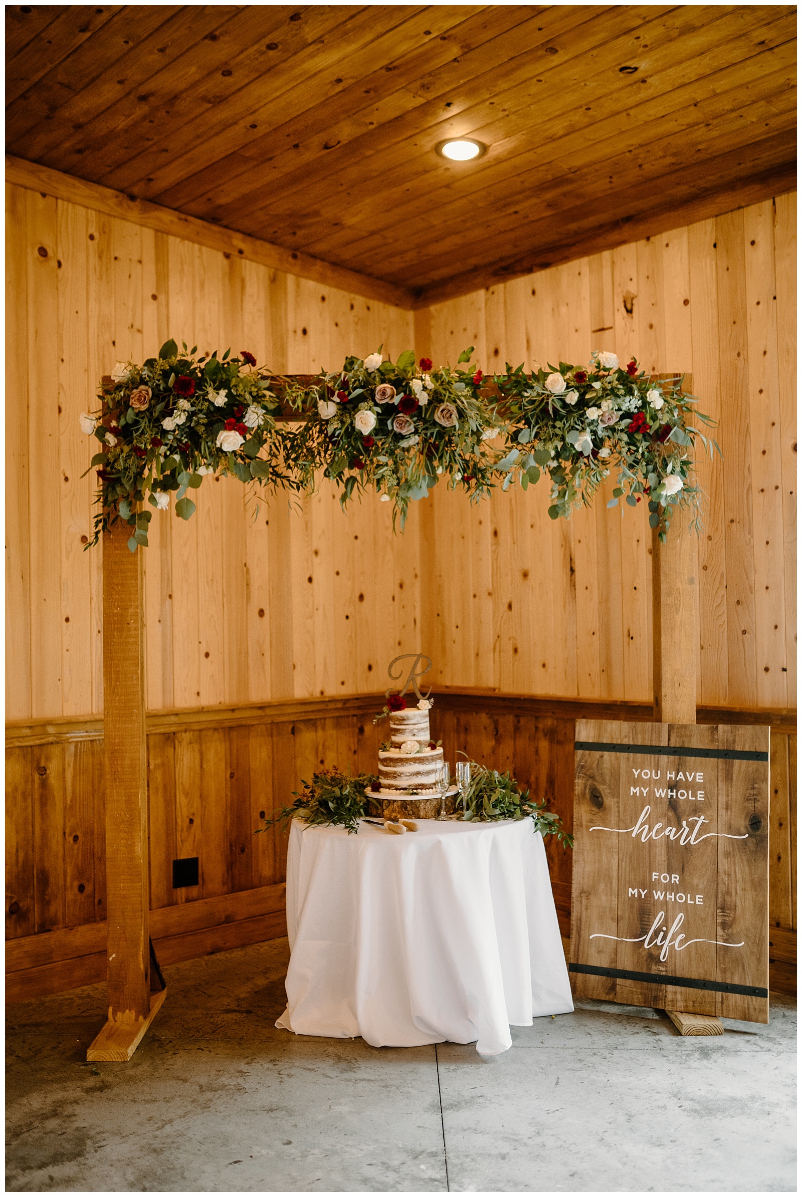 Cake table at wedding reception with floral arch overhead and lots of greenery